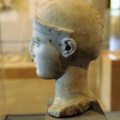 Vers 470-460 av. J.-C.
Fabrication : Egine
Marbre parien, l'ensemble du visage devait être rehaussé par de la peinture. Les cils sont en métal. Le cou venait s'encastrer dans le corps d'une statue
